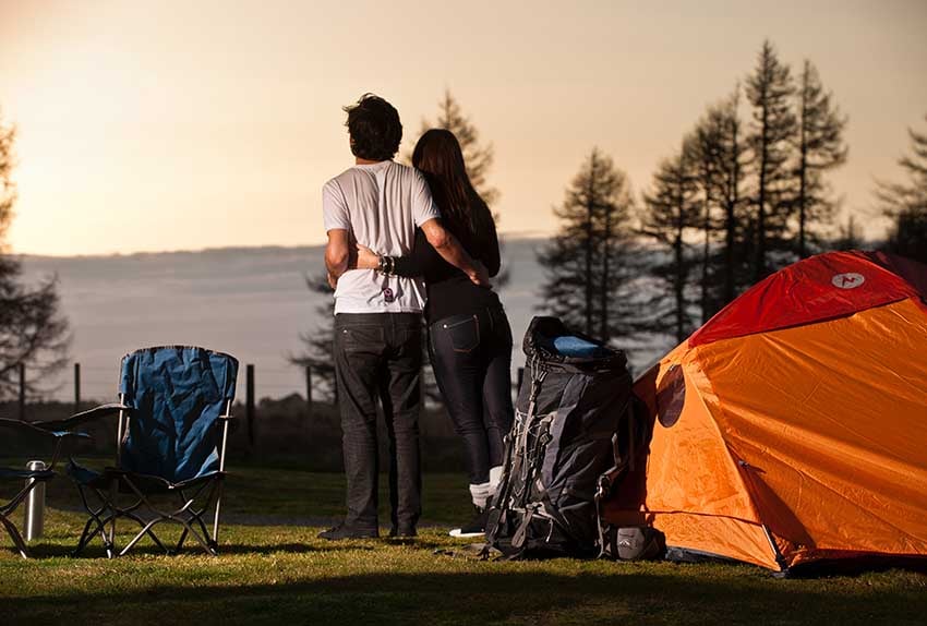 Two campers with their arms around each other next to a marmot tent watching the sun set from Discovery Lodge Tongariro