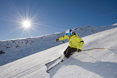 A skier in a yellow jacket skiing down a mountain slope with sun shinning