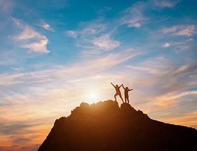 Silhouette of two hikers with arms raised standing on a rocky peak 