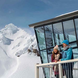 Two people standing outside the Knoll Ridge Cafe with the Pinnacles of Mt Ruapehu in the background at Whakapapa